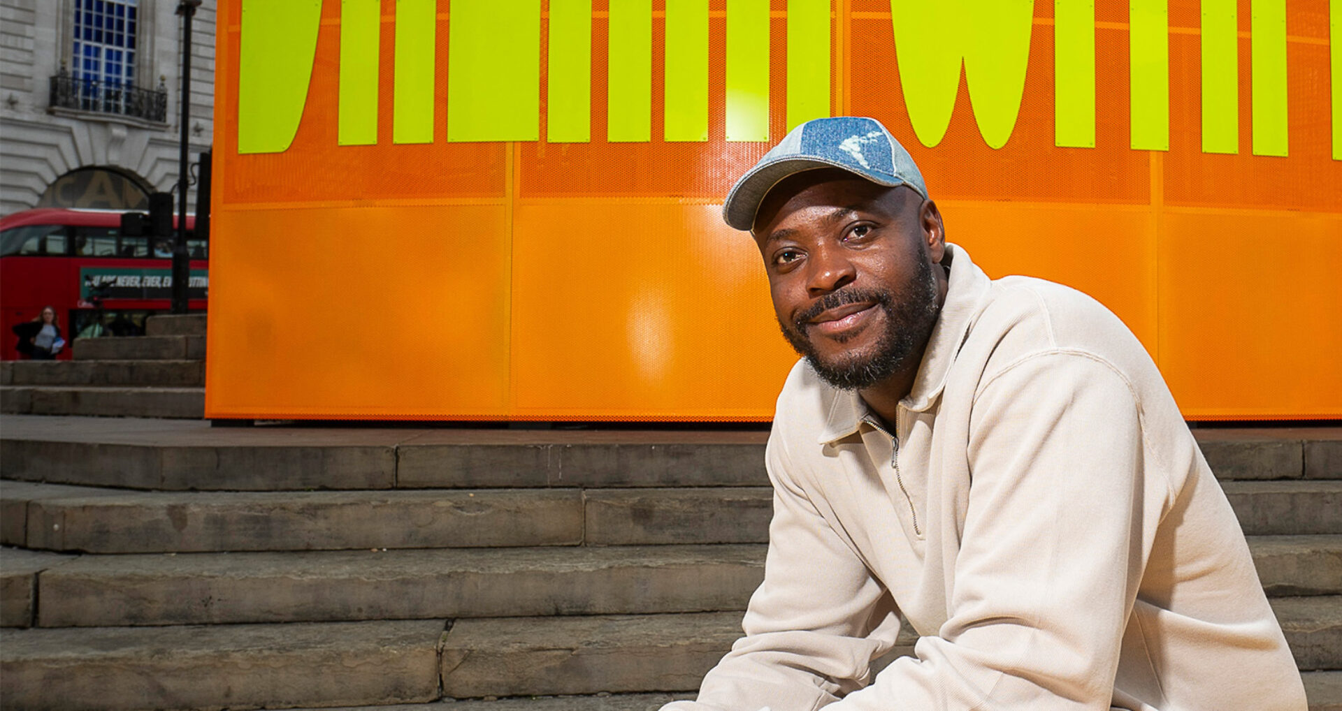 A smiling bearded man in a denim cap and beige pullover sits on steps before a large orange and yellow sign, a red bus behind him.