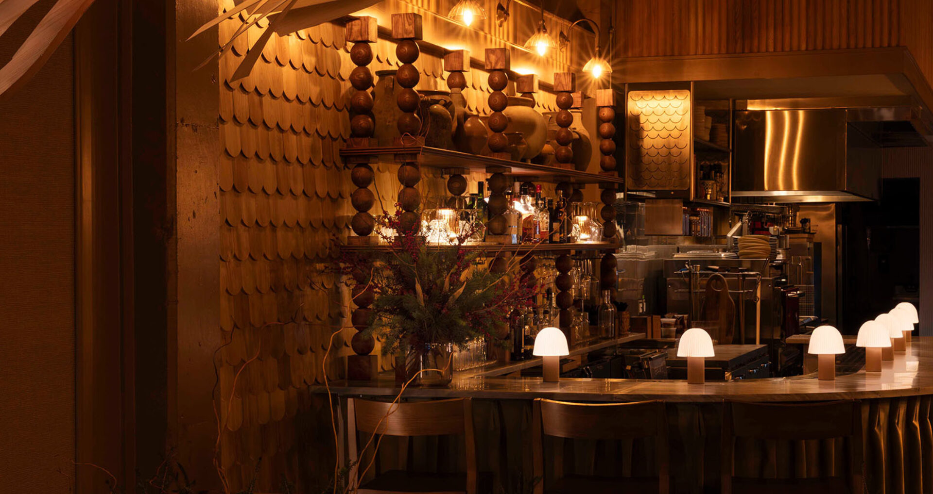 Warm, dim bar with scalloped wooden wall panels, stacked column shelves of vases and bottles, and a curved counter lit by mushroom lamps.