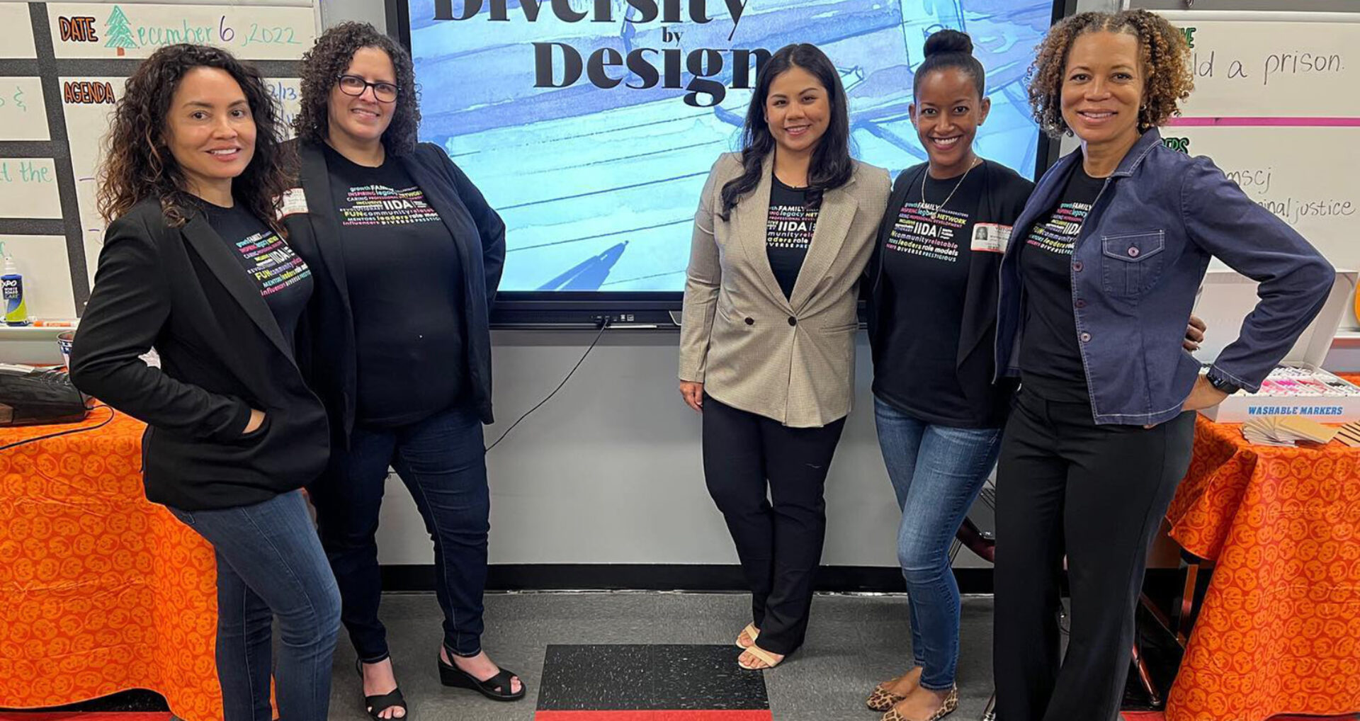 Five women stand together in a classroom, promoting the theme "Diversity by Design" against a backdrop of educational materials.