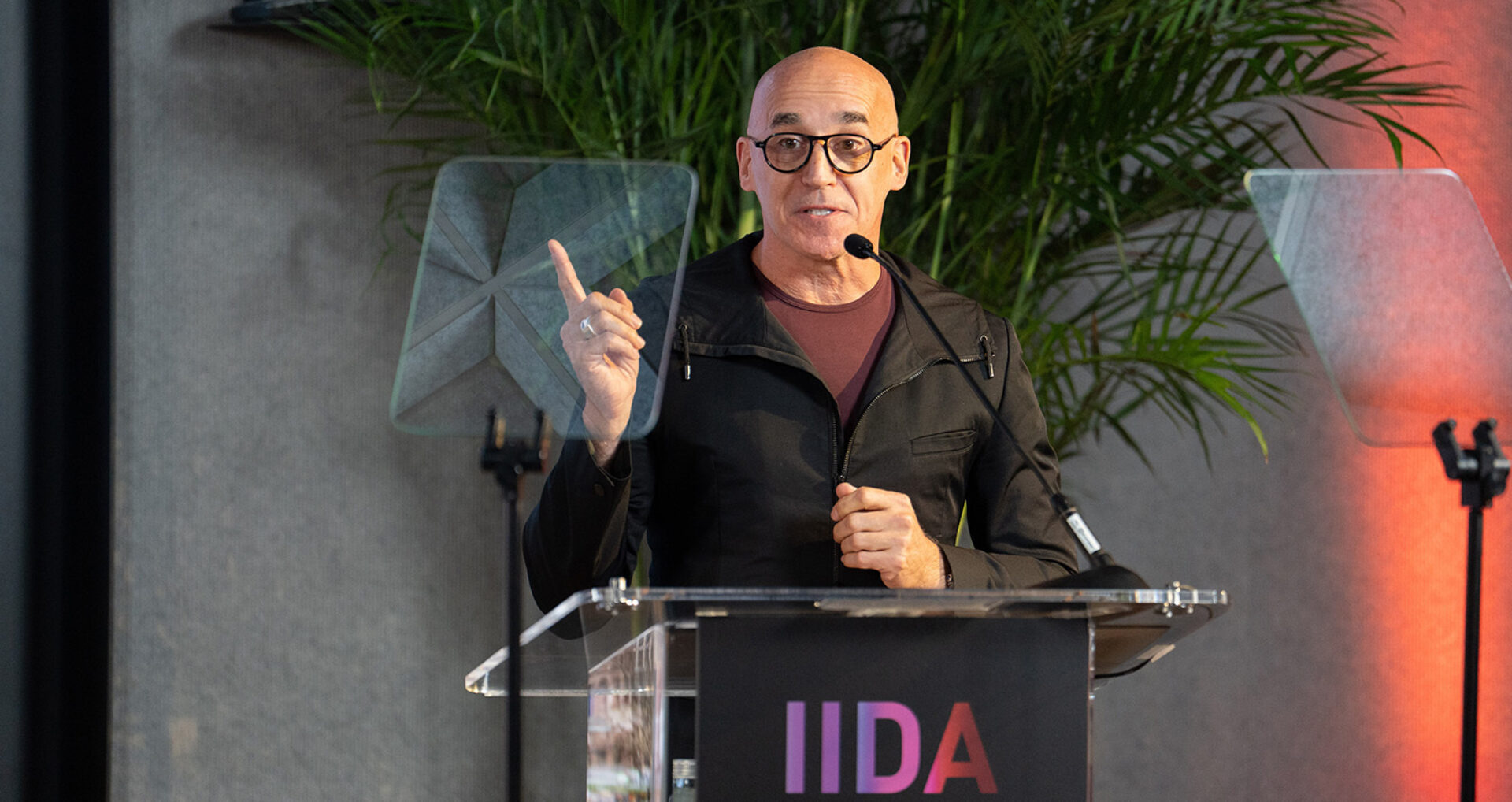 Bald man with round glasses and a shirt under a black jacket speaks into a mic at a clear lectern labeled IIDA, pointing with one finger.