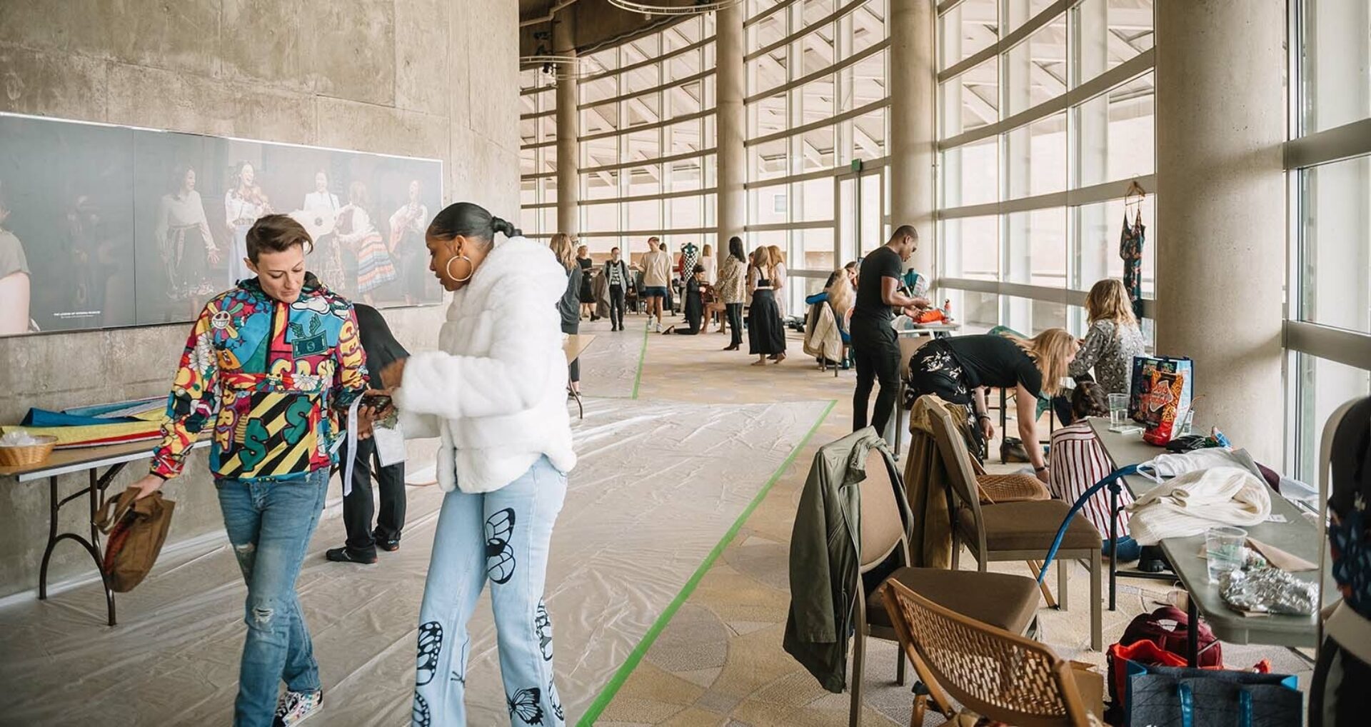 A vibrant gathering space features people interacting, preparing items, and engaging in creative activities amidst large windows and artwork.