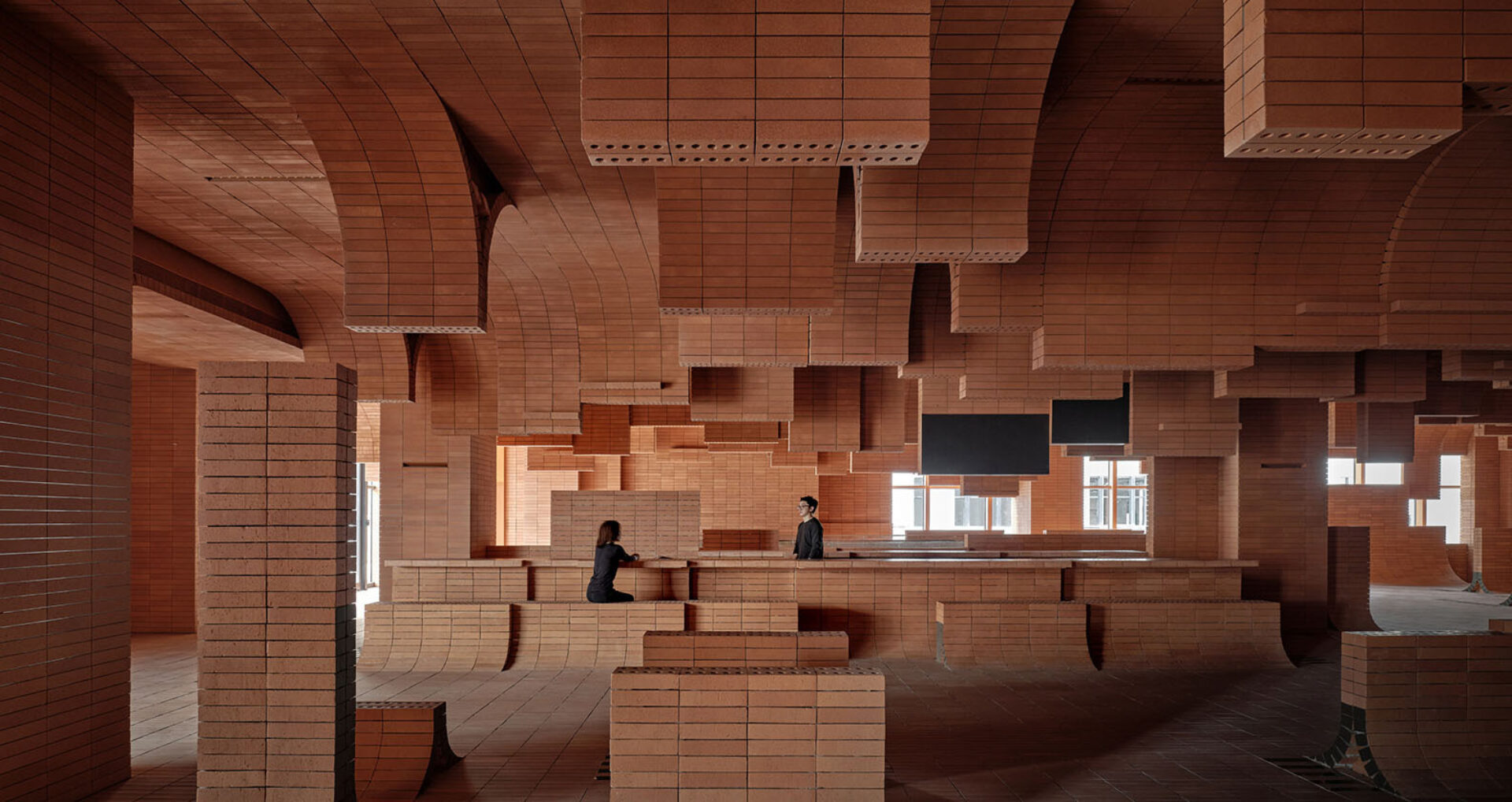 A spacious interior showcases intricate brickwork with arches and geometric shapes, where two people interact amidst the design.