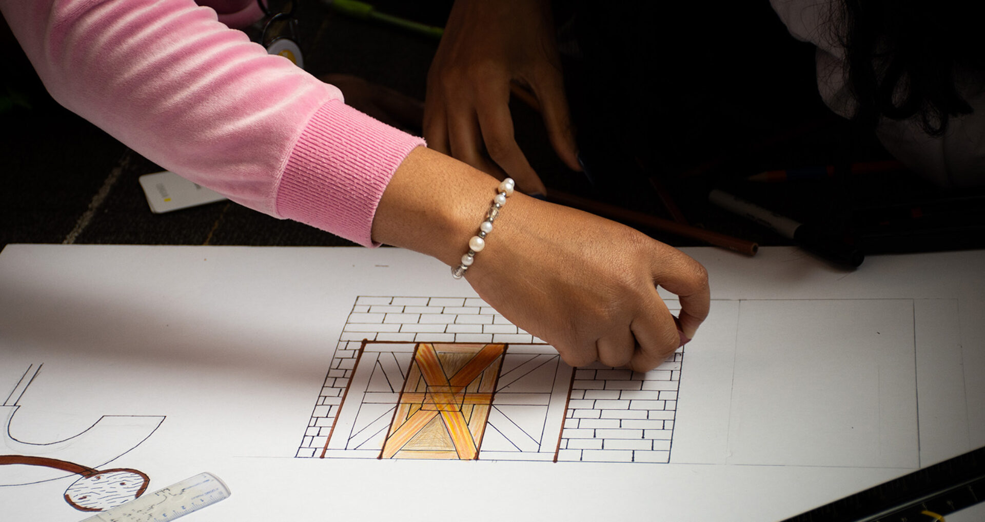 A hand is sketching a detailed architectural design on paper, featuring a window with a wooden frame and brickwork, surrounded by art supplies.