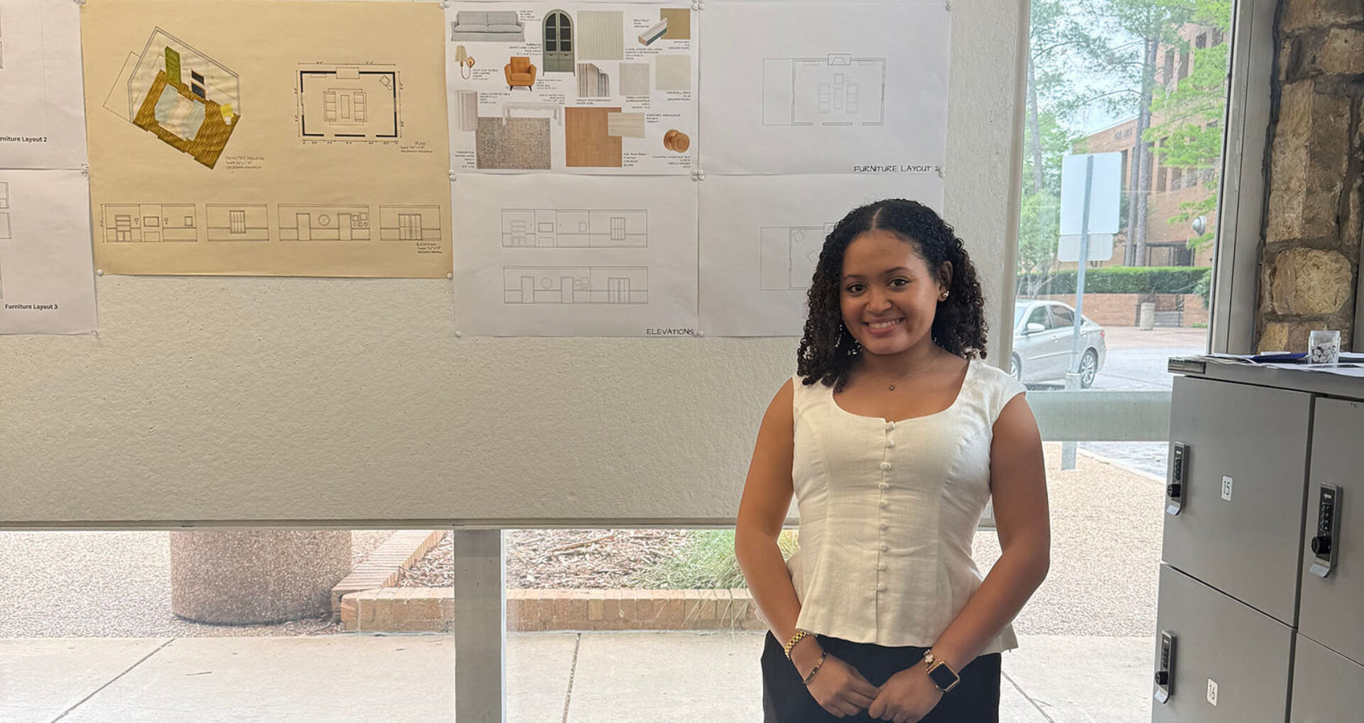 Young woman with curly hair smiles in a white buttoned blouse beside a wall of architectural drawings and lockers near a glass window.