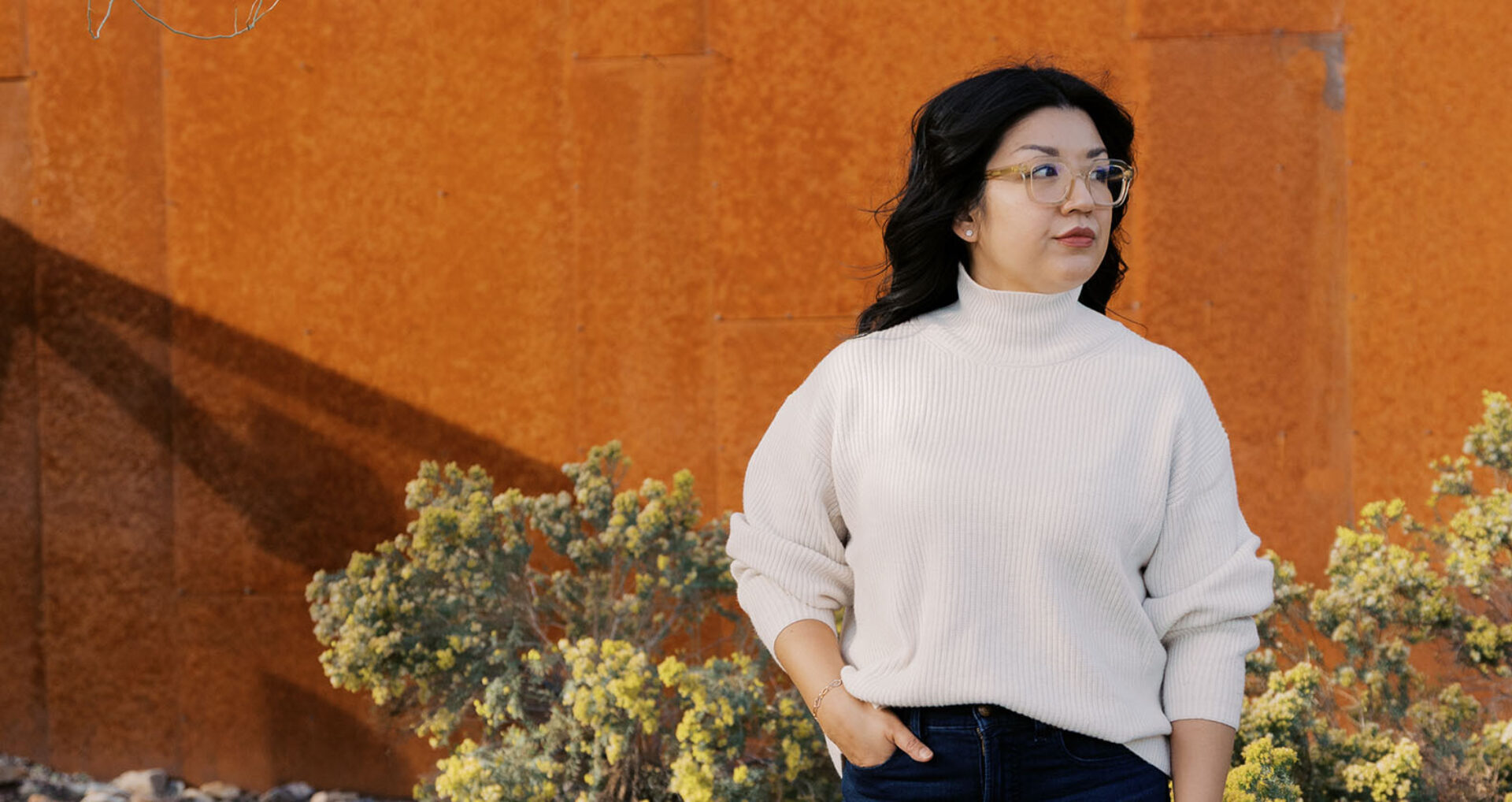 Woman with dark hair, clear glasses and cream turtleneck stands with hand in pocket by a rust wall and yellow shrubs, gazing right.