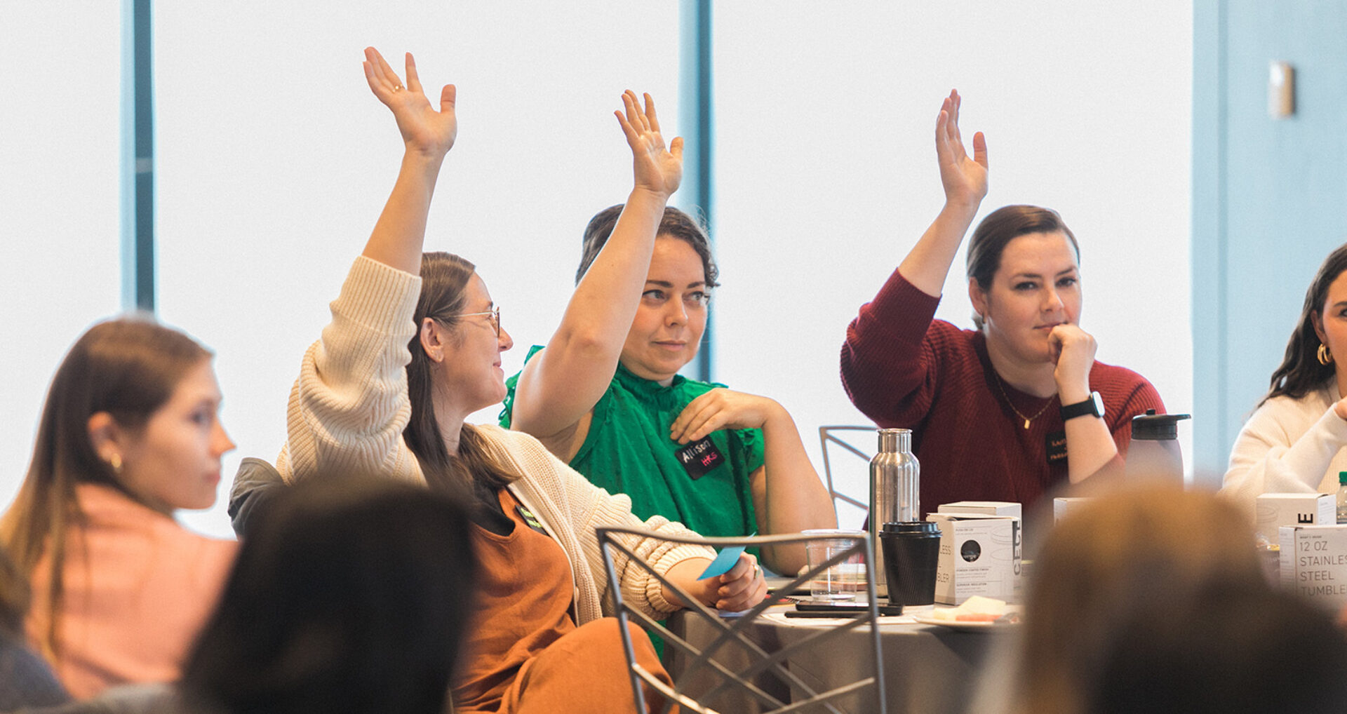 Three women seated at a conference table raising hands, one in a green blouse wearing a name tag reading Allison, cups and notebooks nearby.
