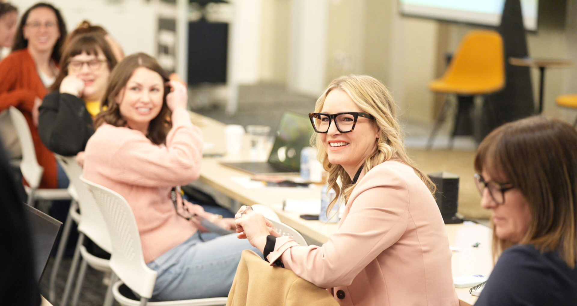 A group of smiling women engage in conversation during a meeting, with a well-lit room and modern furnishings in the background.