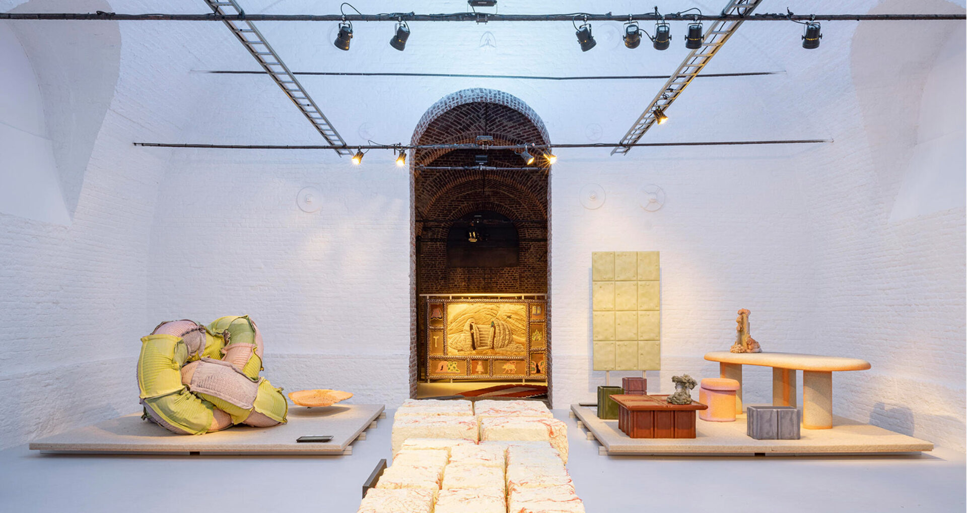 A modern art gallery showcases abstract sculptures and furniture pieces under soft lighting, with textured walls and unique displayed artworks.