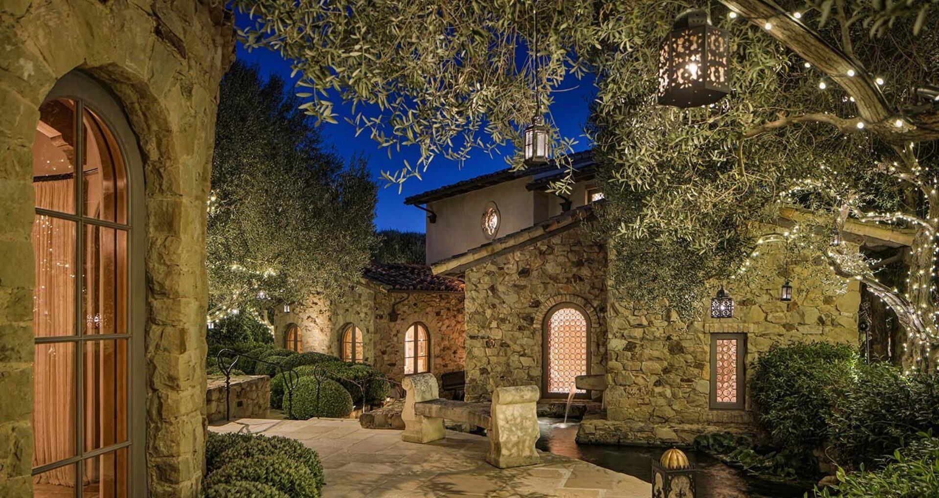 Stone Mediterranean courtyard at twilight with arched windows, lanterns and string lights in olive trees, stone bench by a water channel.