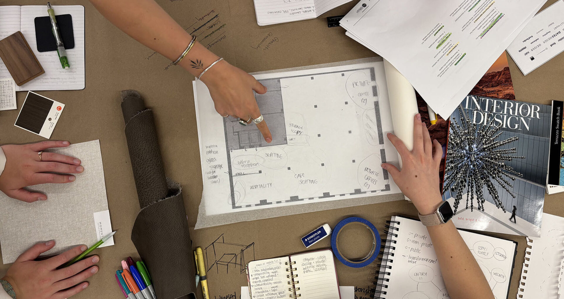 Overhead table with hands pointing at a taped floor plan, surrounded by fabric swatches, leather roll, pens, tape and Interior Design mag.