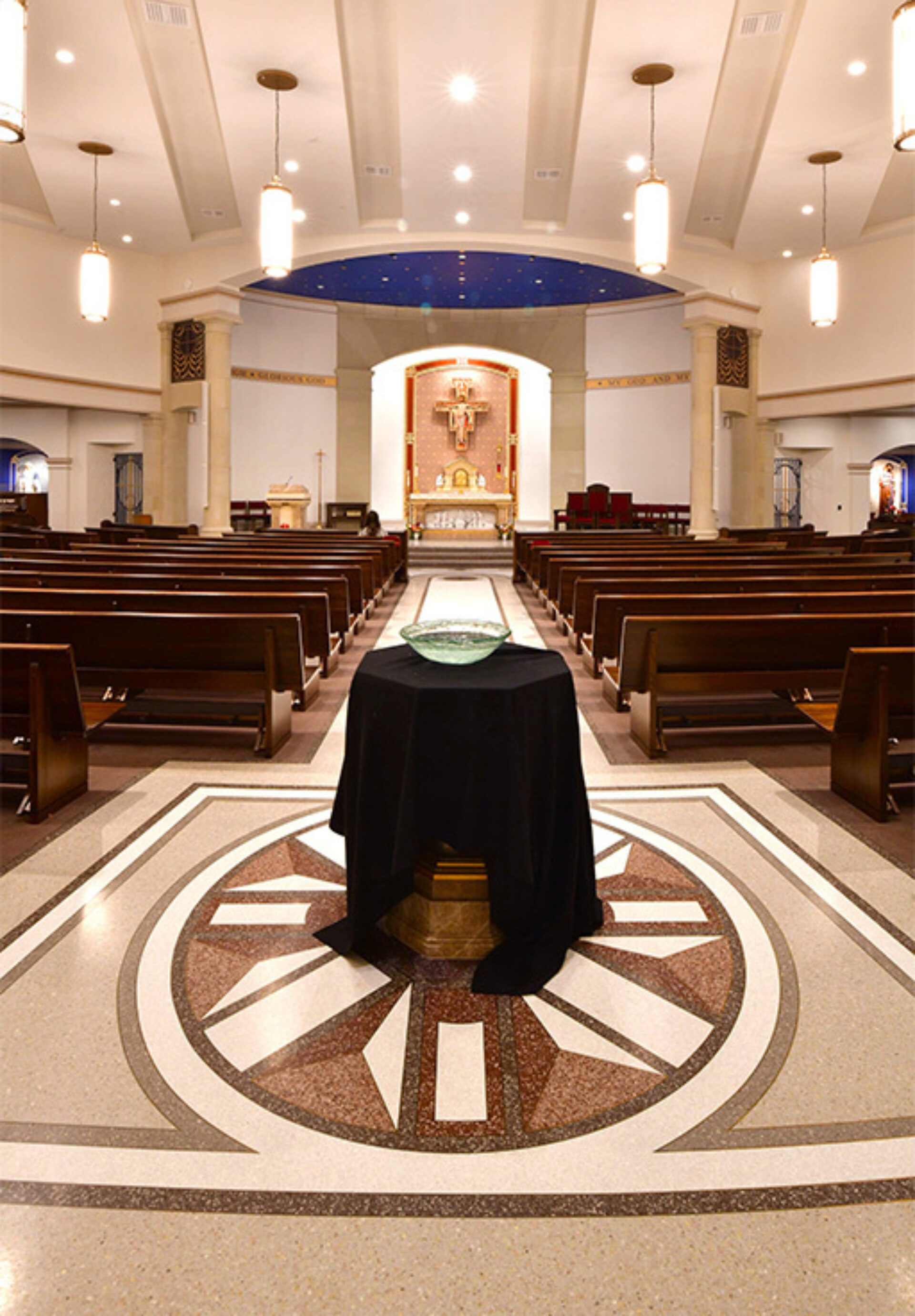 Catholic church interior with central baptismal font draped in black and glass bowl, wooden pews lining the aisle, and altar crucifix.