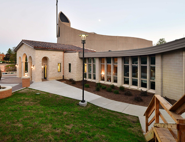 Curved-roof church at dusk with arched stone entry, rows of tall windows, bronze statue by a circular fountain, lamp post and wooden steps.