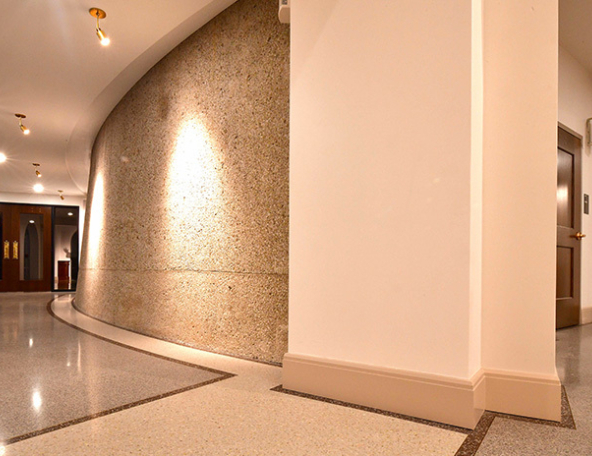 Wide curved interior corridor with terrazzo floor, textured concrete wall lit by recessed spotlights, and wooden doors at the end.