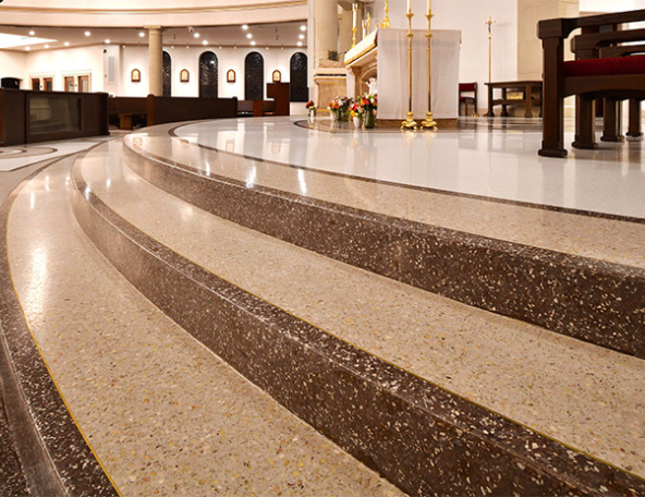 Curved terrazzo steps leading up to a bright sanctuary altar with brass candlesticks, floral bouquets, wooden pews and polished marble floor.