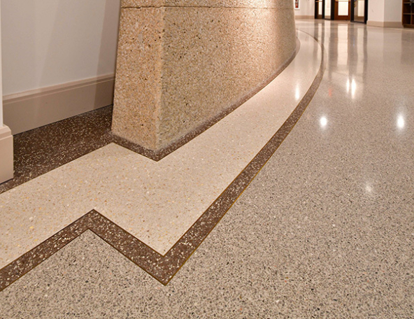 Polished terrazzo corridor floor with beige curved wall and brown zigzag inlay leading toward distant doors, glossy light reflections.