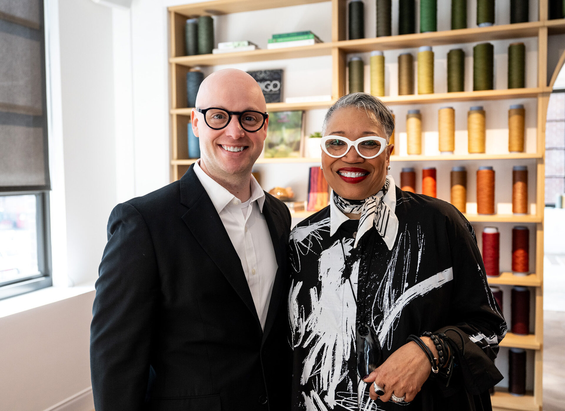 Smiling bald man in black suit beside woman with white cat-eye glasses, red lipstick and black patterned blouse; thread spools on shelves.