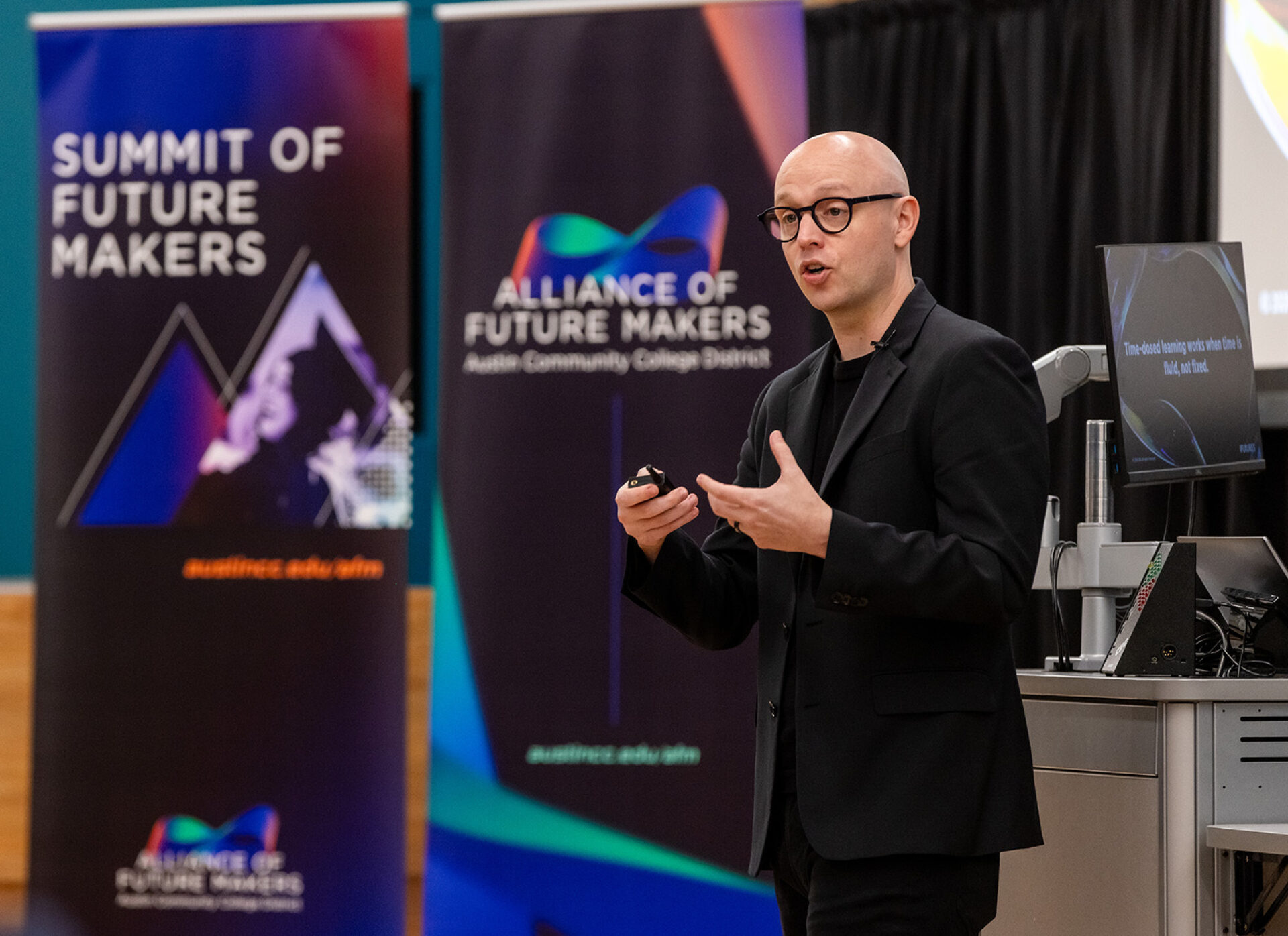 Speaker with bald head and glasses in black suit gestures while holding a clicker, framed by Summit of Future Makers and Alliance banners.