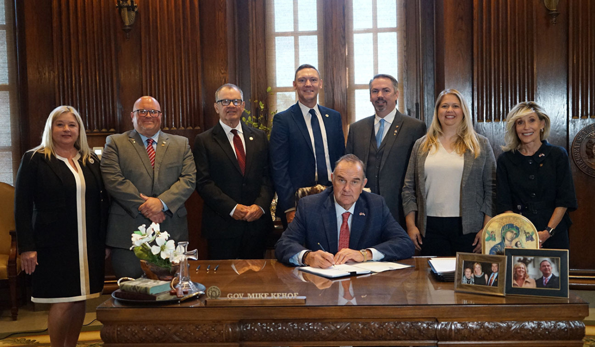 Seated man signs documents at an ornate wooden desk in a wood-paneled office while seven formally dressed colleagues stand behind him.