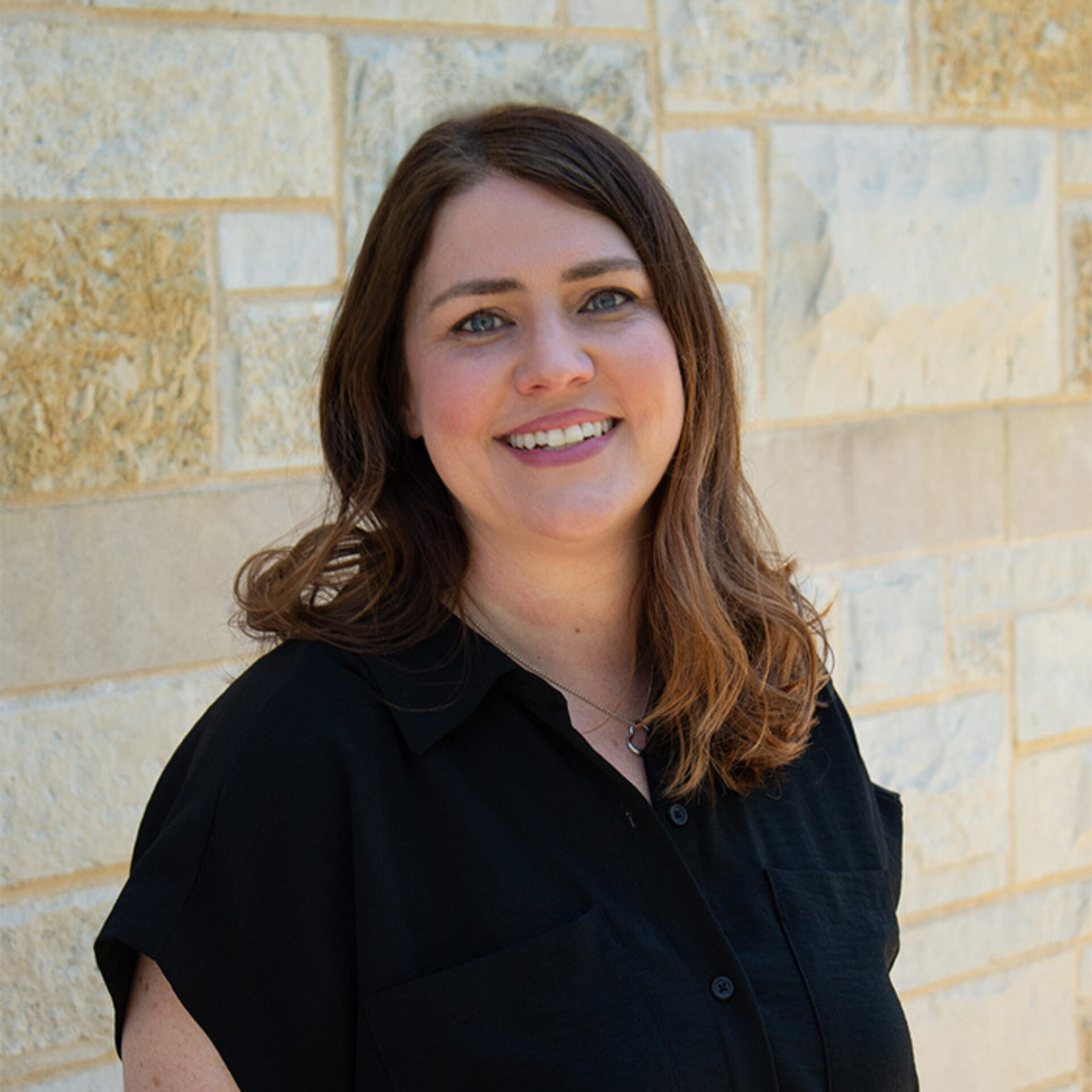 Smiling woman with shoulder-length brown hair wearing a black blouse and small pendant necklace, standing against textured light stone wall.