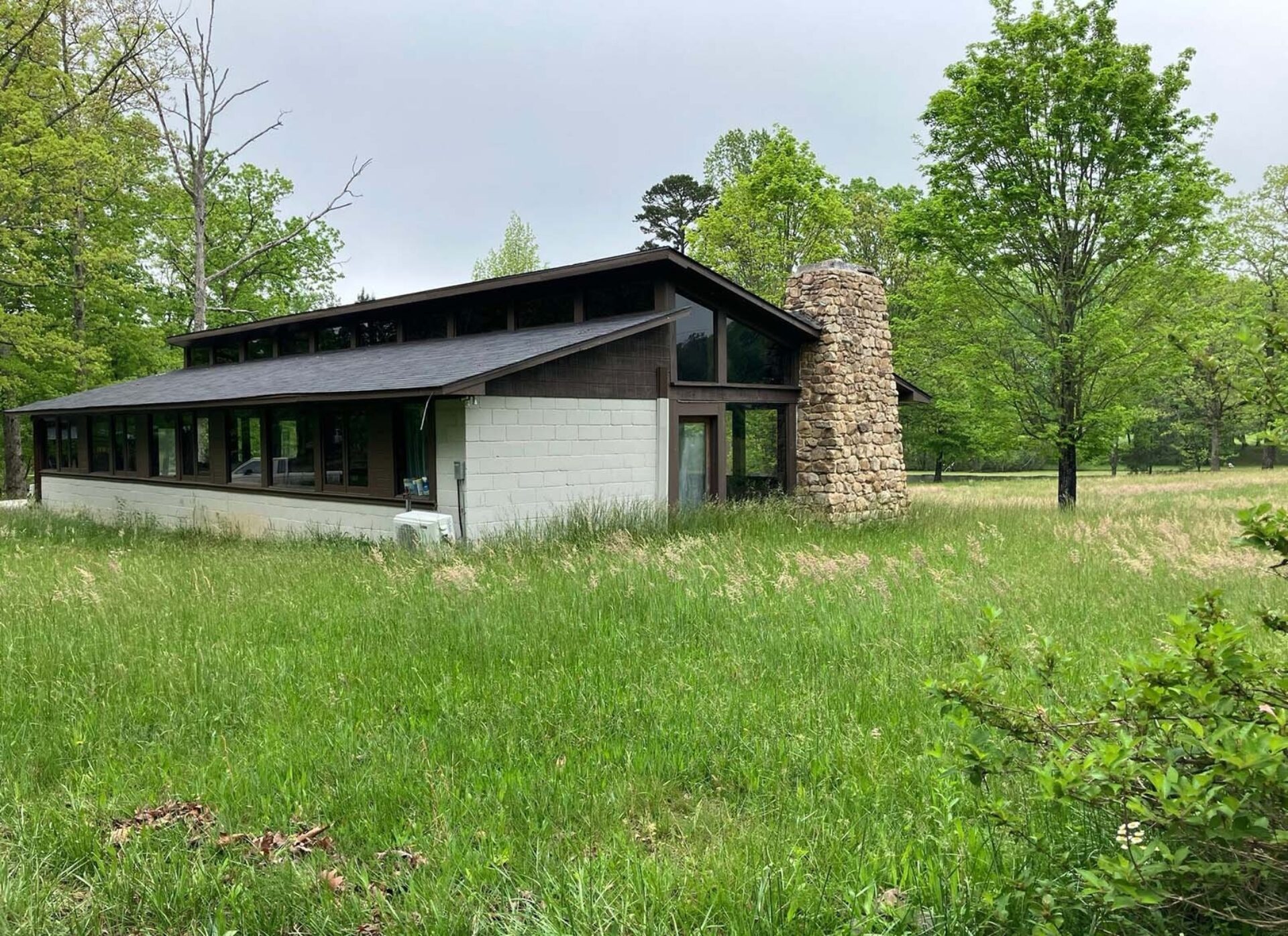 A modern-style building surrounded by tall grass and trees, featuring a prominent stone chimney and large windows for ample natural light.
