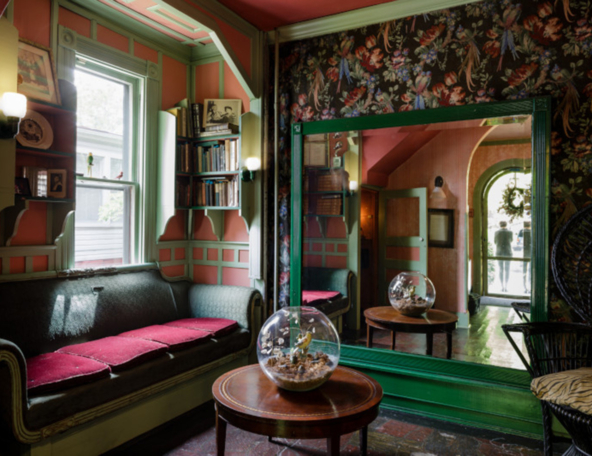 A cozy room featuring a green-framed mirror, vintage bookshelves, a floral wallpaper, and a round coffee table with a glass terrarium.