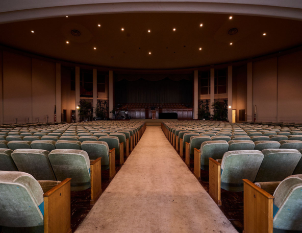 An empty auditorium features rows of worn green chairs leading to a stage, illuminated by warm overhead lights, evoking a nostalgic atmosphere.
