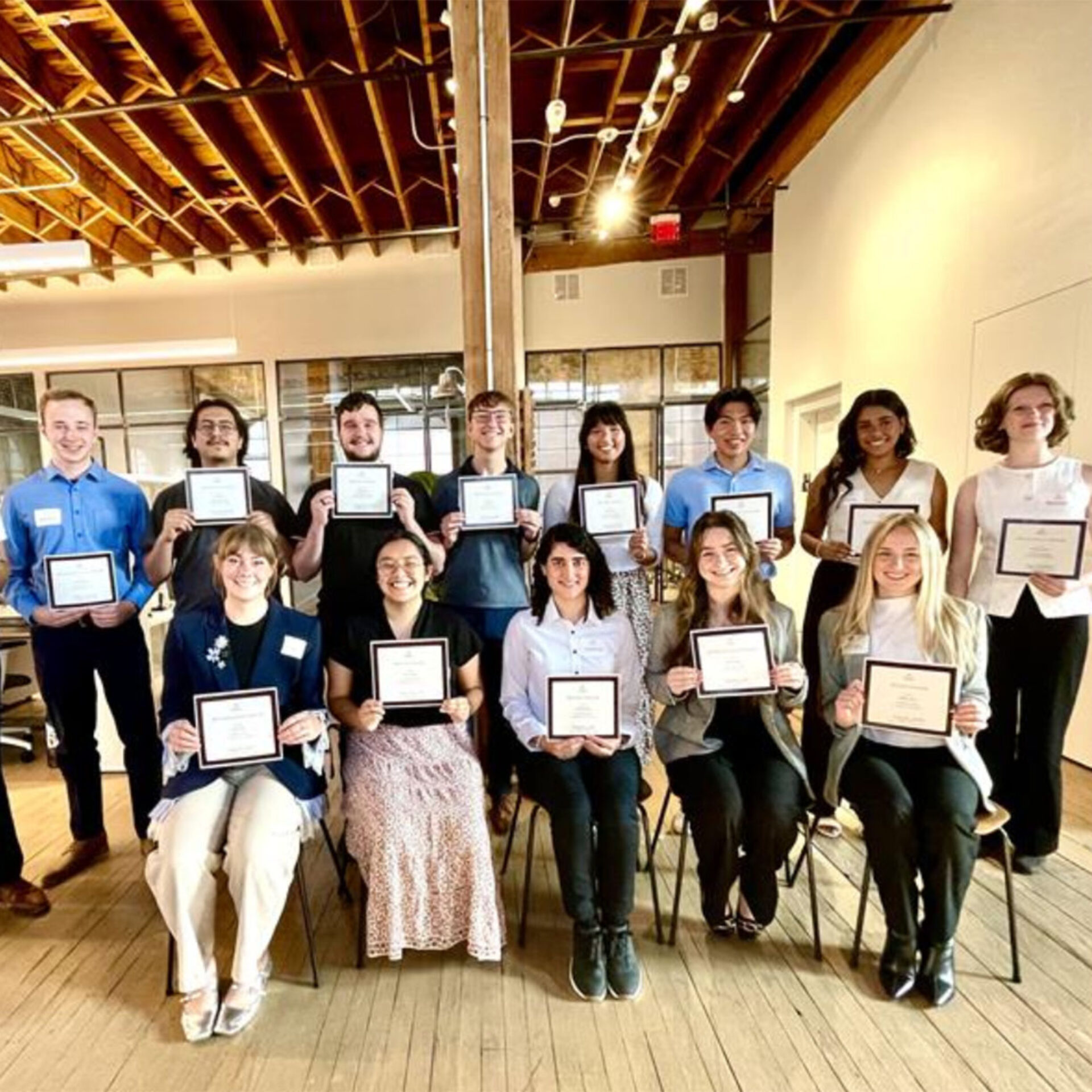 Fifteen smiling young adults in two rows hold framed certificates inside a sunlit loft with exposed wooden beams and hardwood floors.