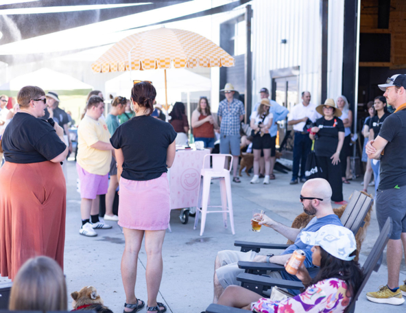 Outdoor festival crowd gathers around a yellow checkered umbrella as a woman speaks into a microphone and attendees sip drinks.