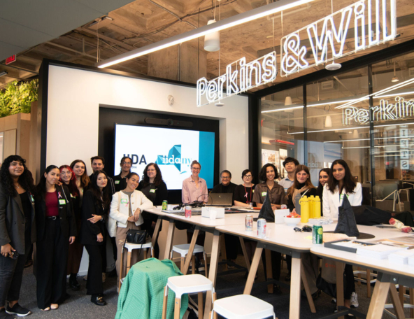 A diverse group of young designers and interns pose around a long table in a Perkins&Will office with presentation screen and neon sign.