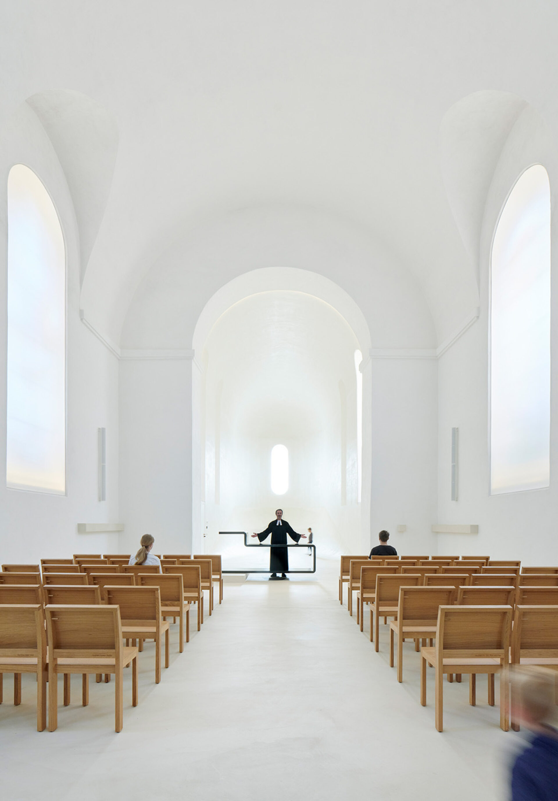 Minimalist white chapel interior with high arched ceiling, wooden pews in rows, a robed minister at the altar and two seated parishioners.
