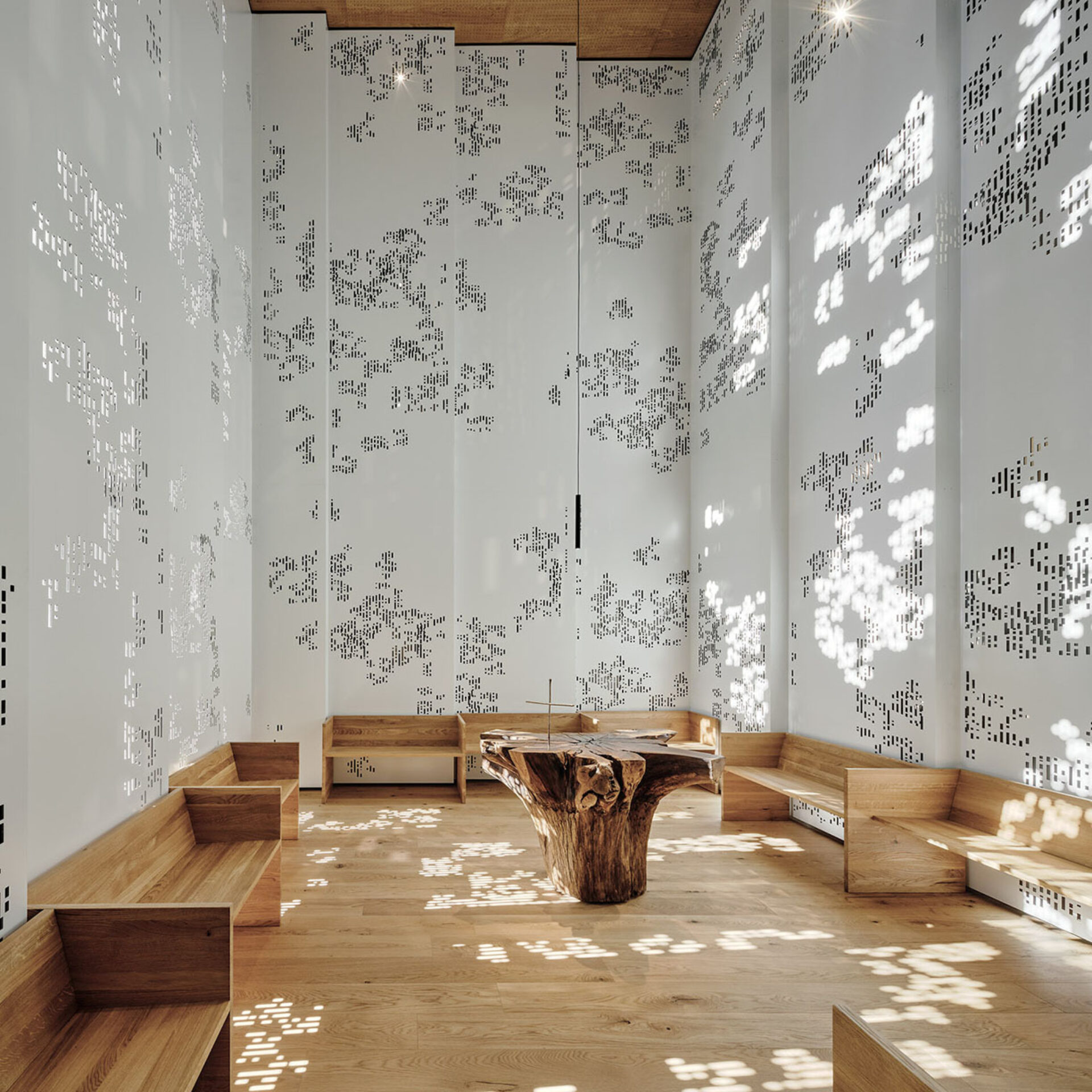 Tall white perforated walls cast dappled light over a wooden floor, perimeter oak benches and a central raw tree-trunk altar table.