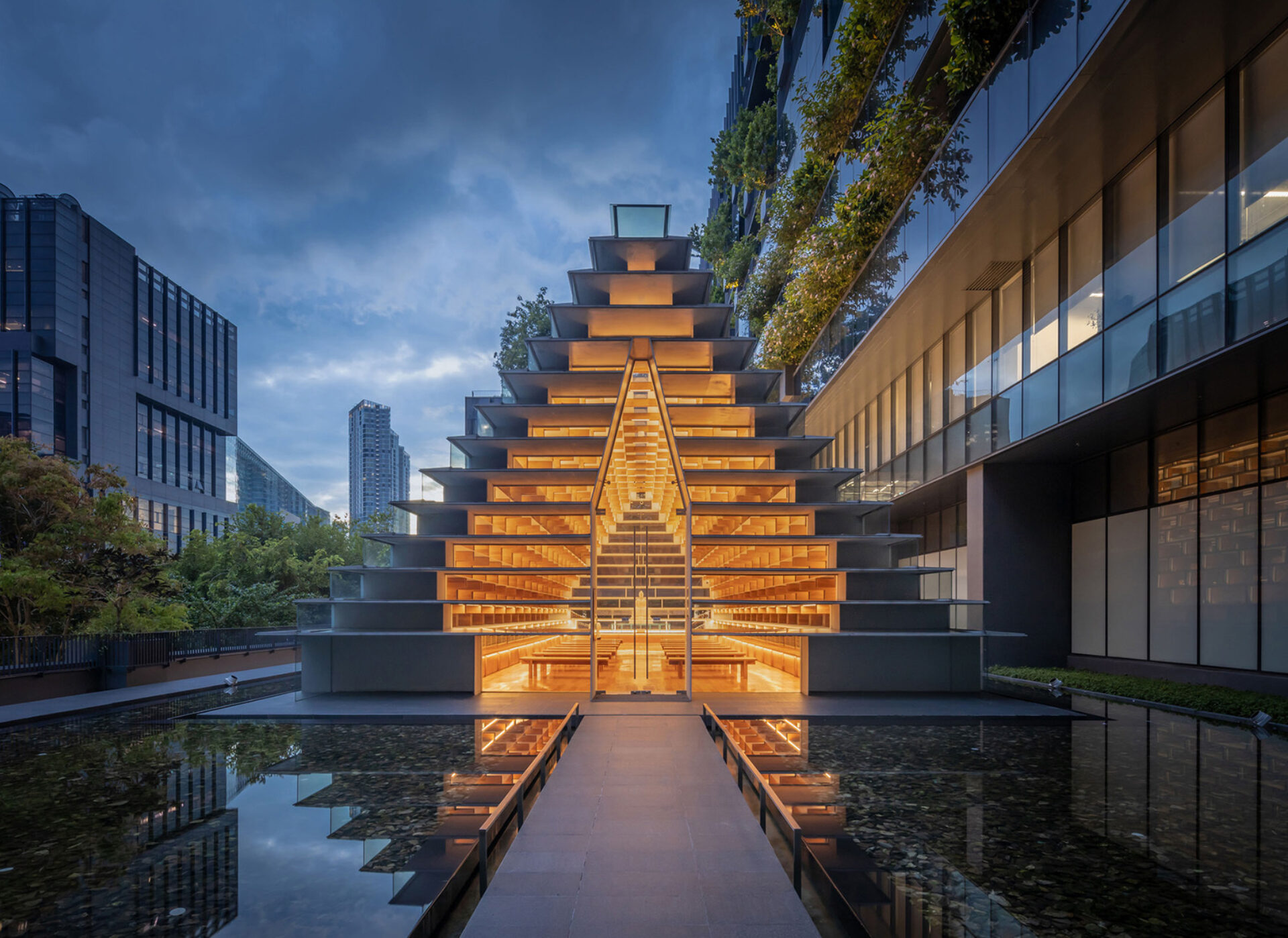A lit stepped wooden pavilion with symmetrical tiers and central glass entrance, reflected in a still pool between glass buildings.