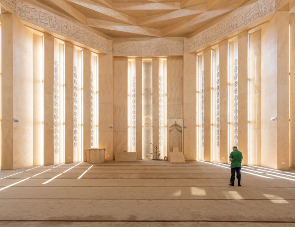 Spacious beige mosque interior with tall sunlit windows, Arabic calligraphy frieze, mihrab, minbar and lone man in green shirt.
