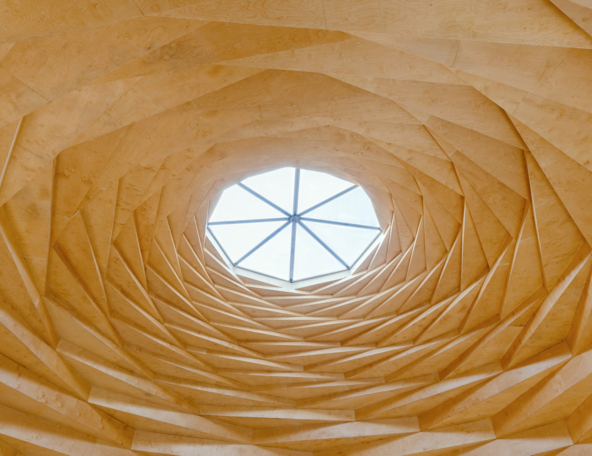Honey-colored wooden panels spiral upward in concentric geometric folds toward an octagonal glass skylight with metal radial spokes.