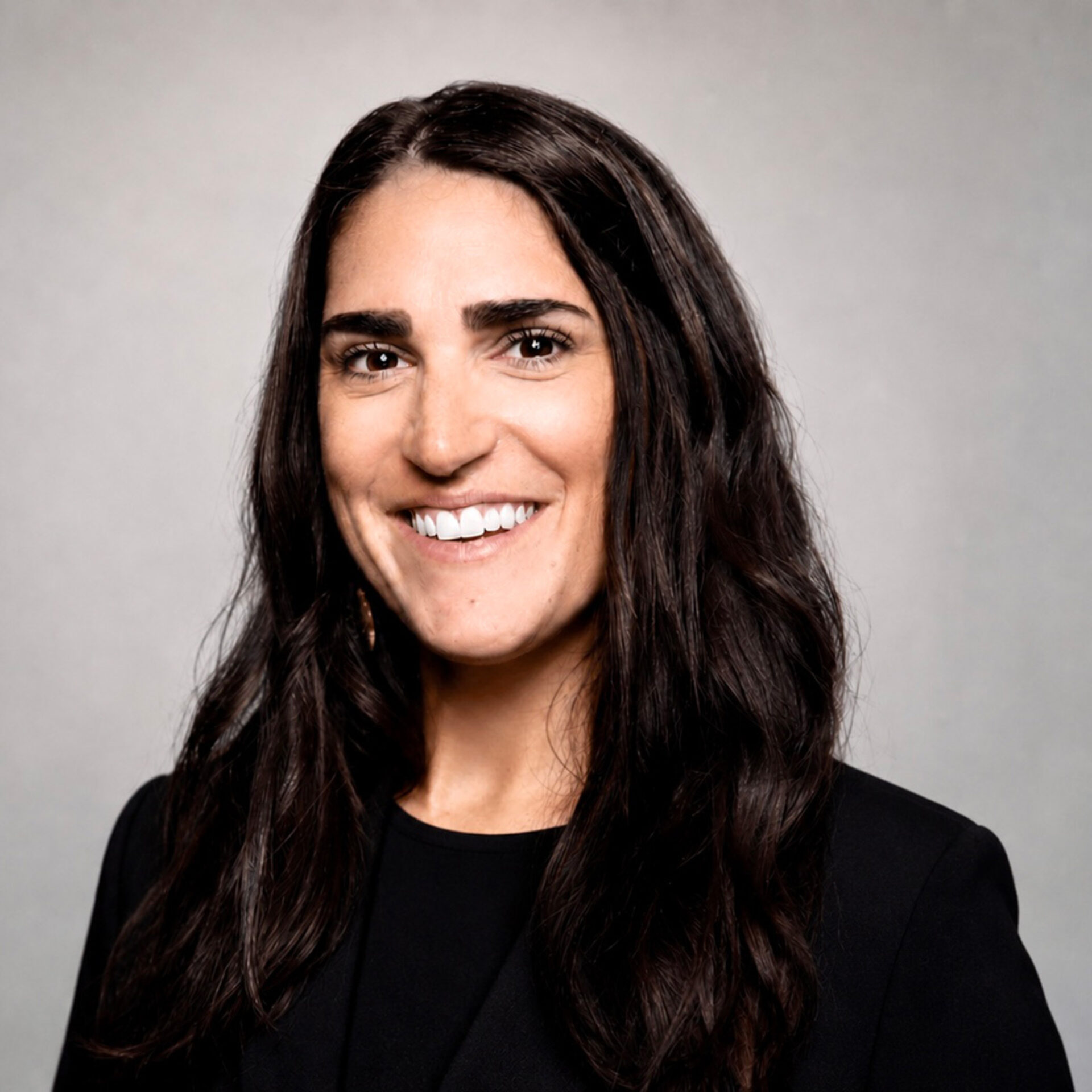 Smiling woman with long dark wavy hair, thick eyebrows and bright white teeth wearing a black blazer against a light gray backdrop.