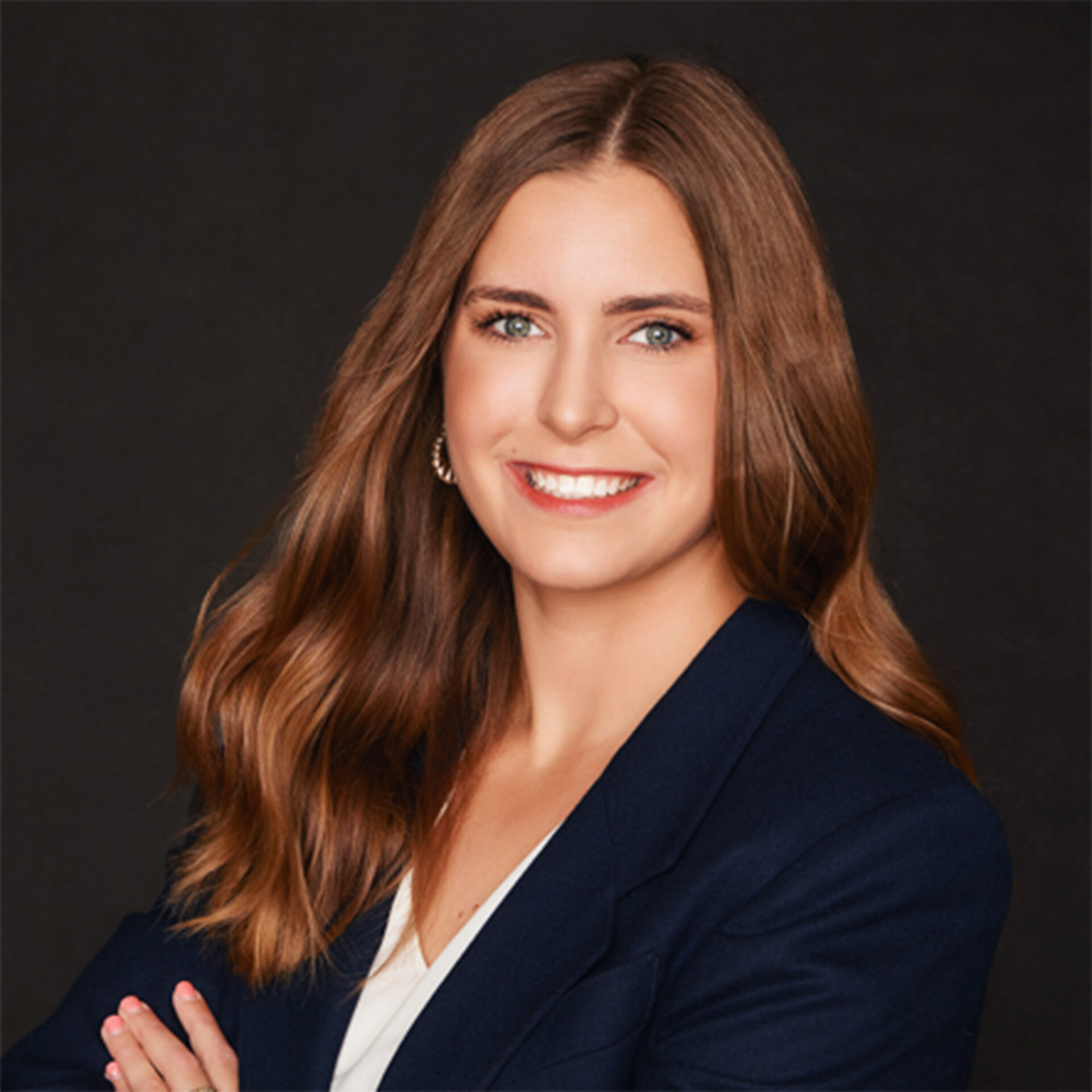 Smiling professional woman with long wavy brown hair, light green eyes, wearing a navy blazer and white blouse, posed against dark backdrop.