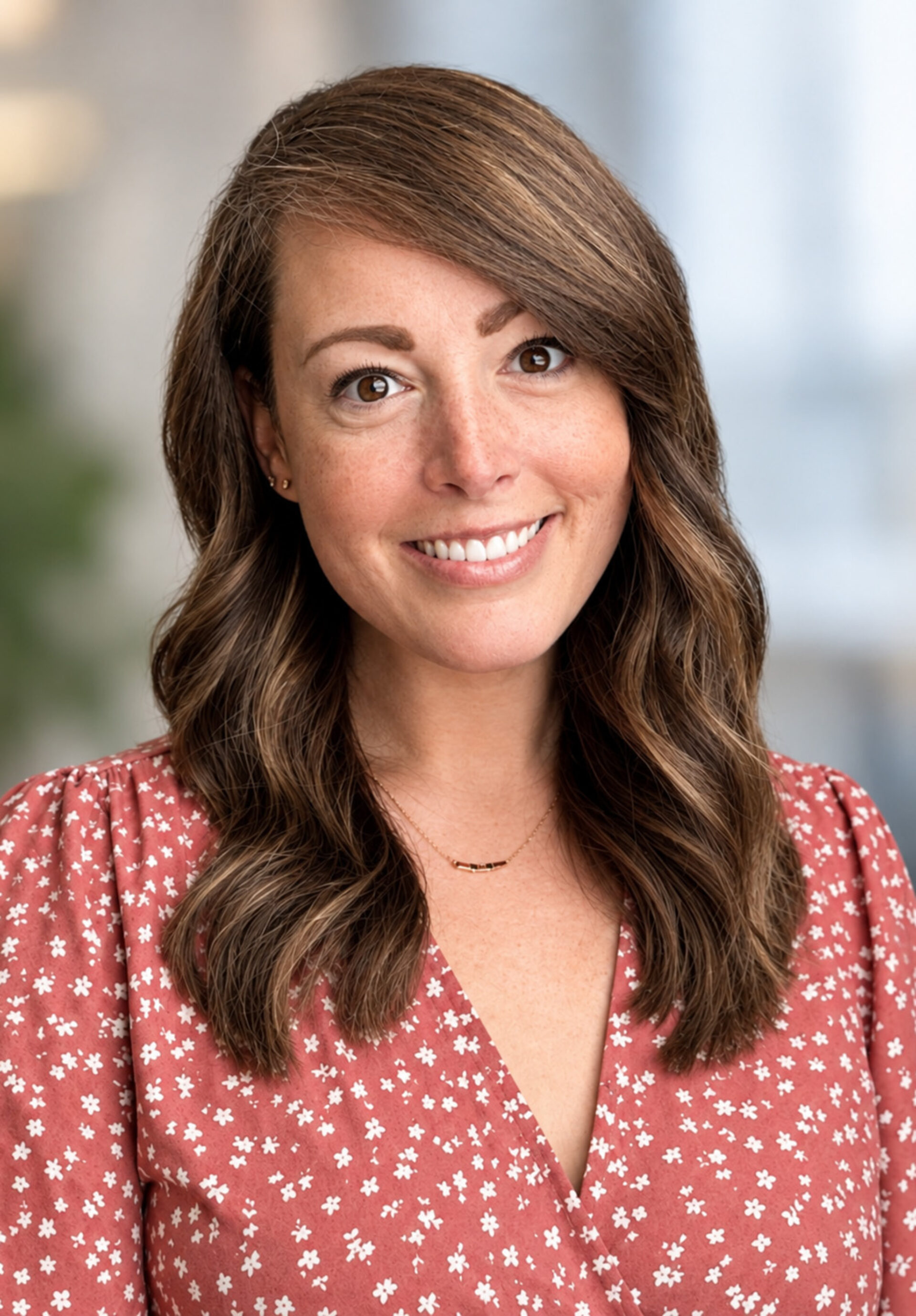 Smiling brown-eyed woman with shoulder-length wavy hair, freckles and necklace, wearing rust floral blouse against soft blurred background.