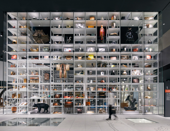 A tall showcase filled with diverse artifacts, including pottery, textiles, and sculptures, with a person walking in front.