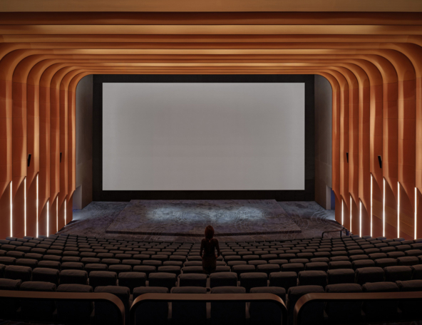 A modern cinema interior featuring an empty auditorium, orange wood accents, a large blank screen, and plush seating.