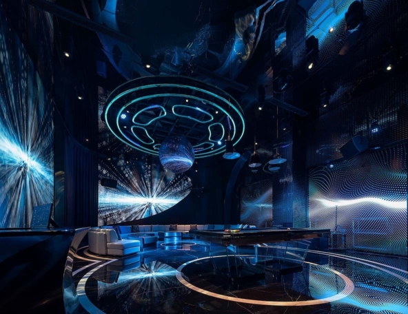 A futuristic lounge illuminated with blue lights, featuring sleek modern furniture, a reflective floor, and abstract visual designs on the walls.