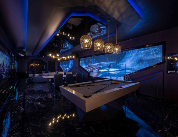 A modern entertainment room features a sleek billiards table, stylish seating, and large screens with dynamic lighting and artwork.