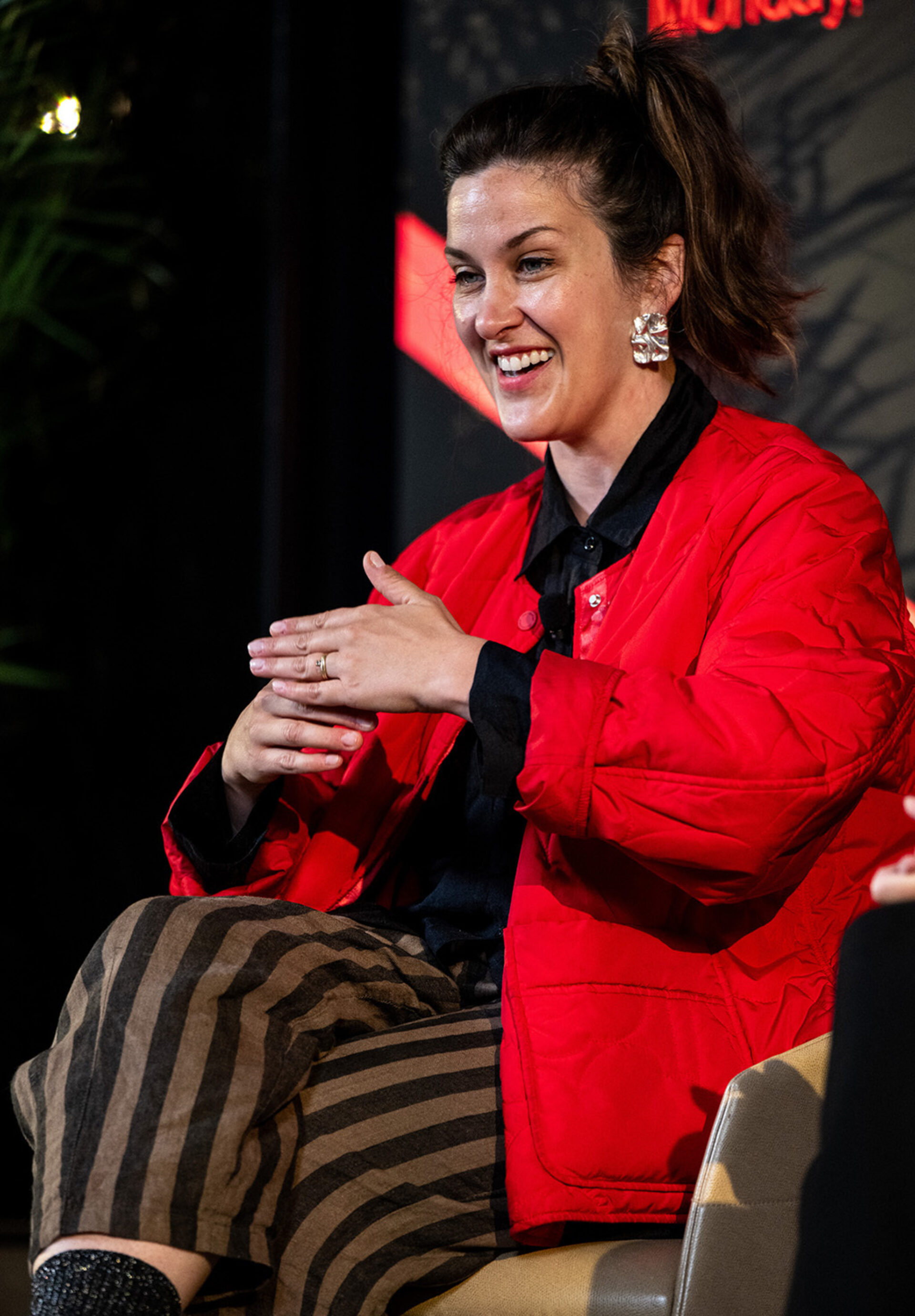 Smiling woman with ponytail and large silver earrings gestures while speaking on a panel, wearing a red quilted jacket and striped pants.