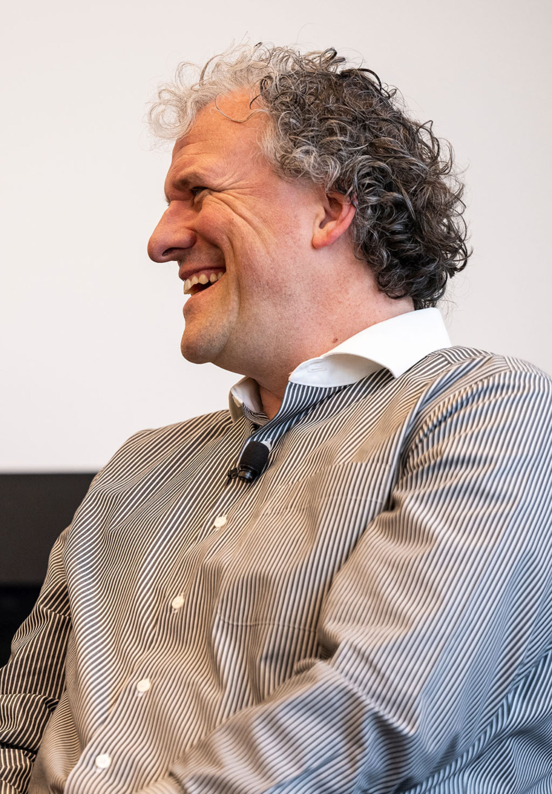 Curly-haired middle-aged man in a striped button-down shirt laughs in profile with a clip-on microphone at his chest, eyes crinkled.