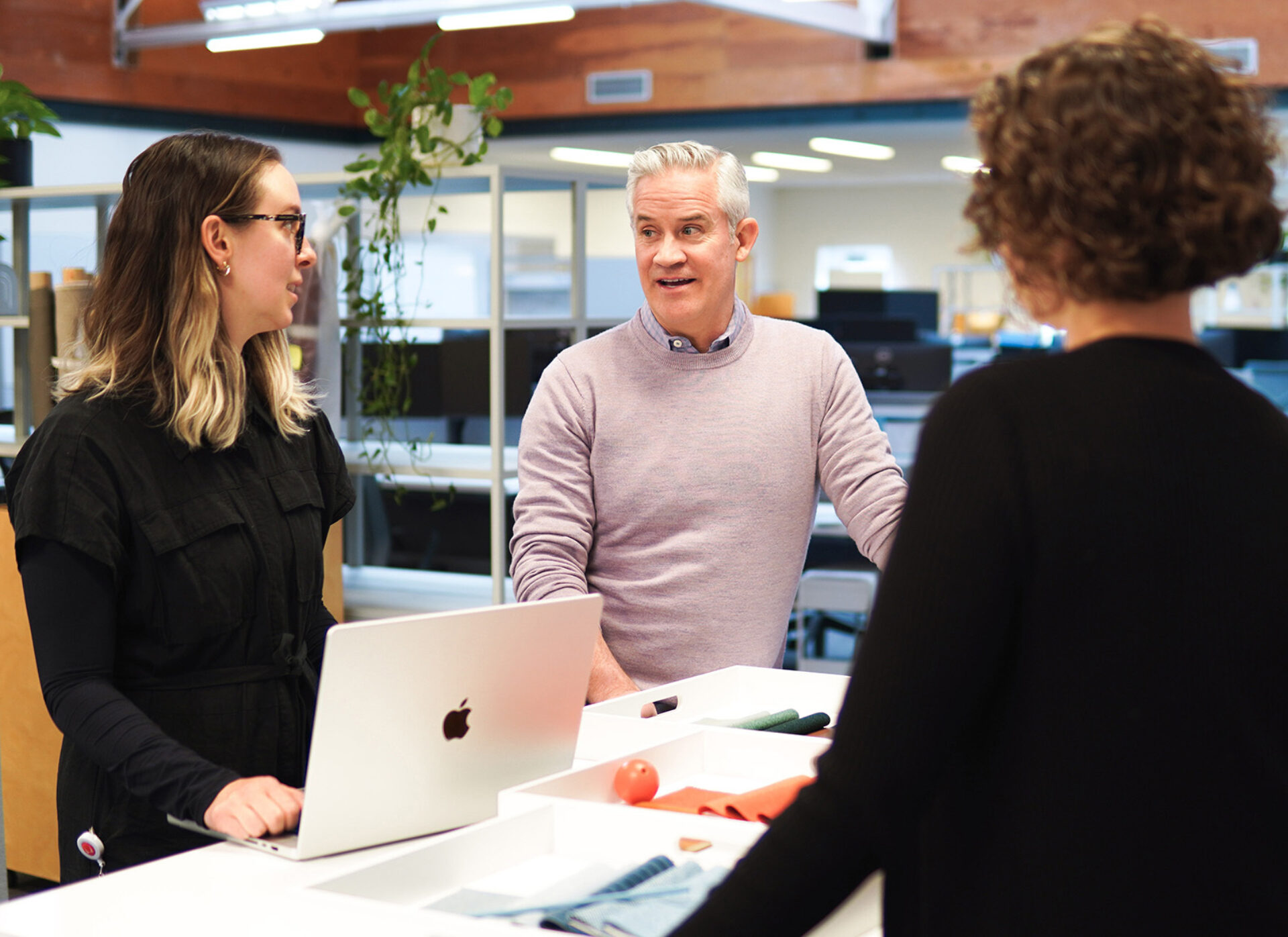3 colleagues stand around a white workspace as a gray-haired man in a lavender sweater speaks while a bespectacled woman types on MacBook.