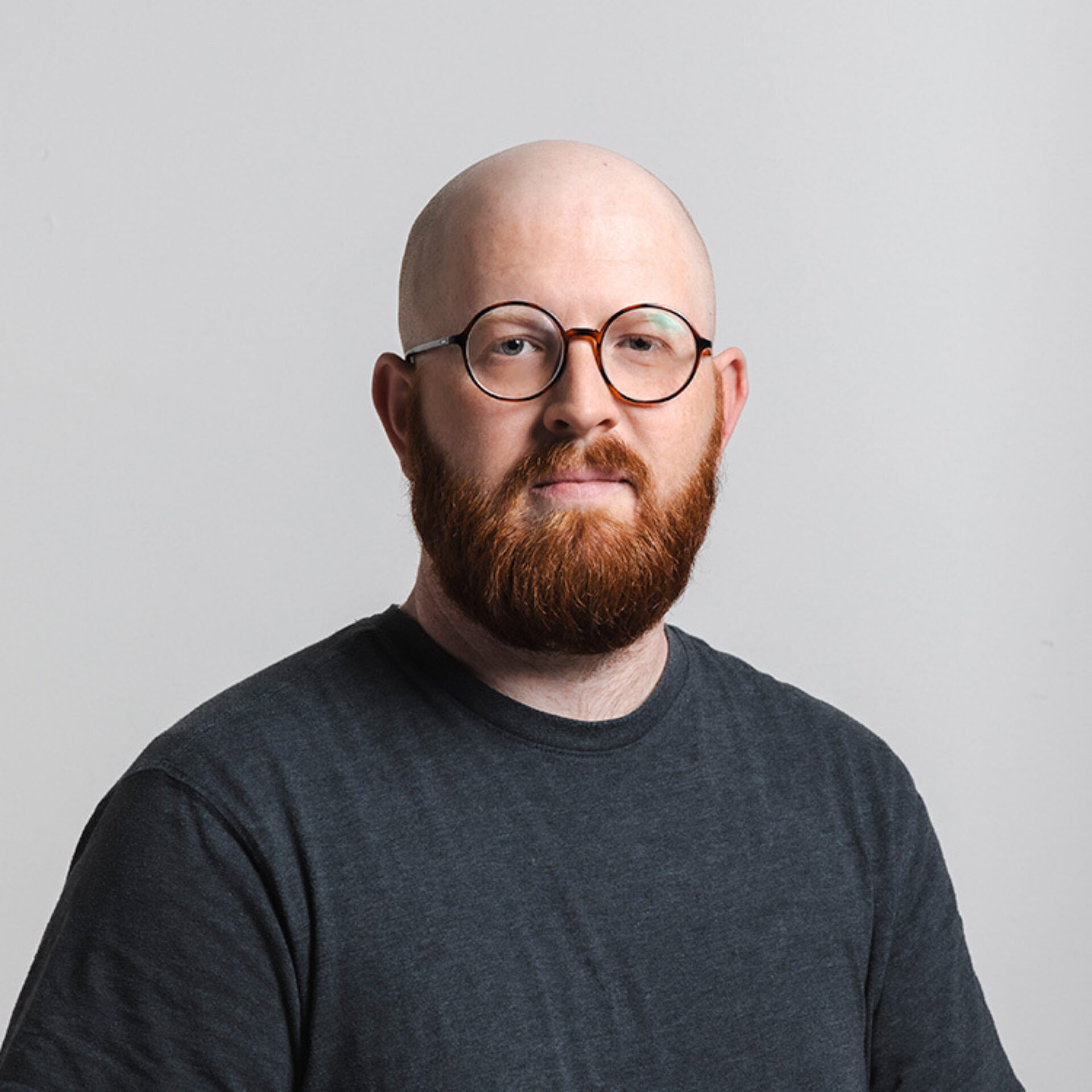 Bald man with round tortoiseshell glasses and full red beard, wearing a dark gray T-shirt, facing camera against a light gray background.