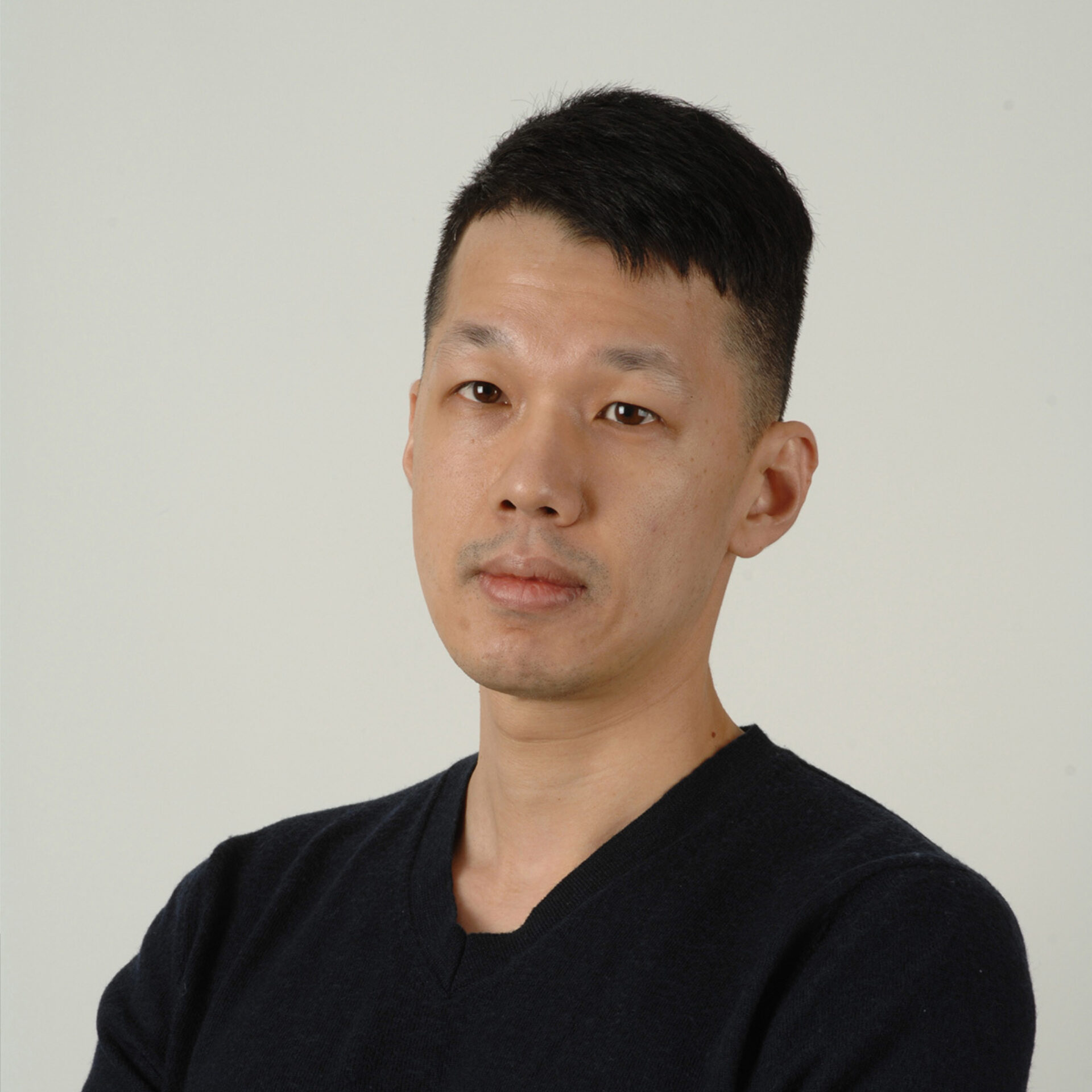 Head-and-shoulders portrait of a man with short black hair, neutral expression, wearing a dark V-neck sweater against a pale background.