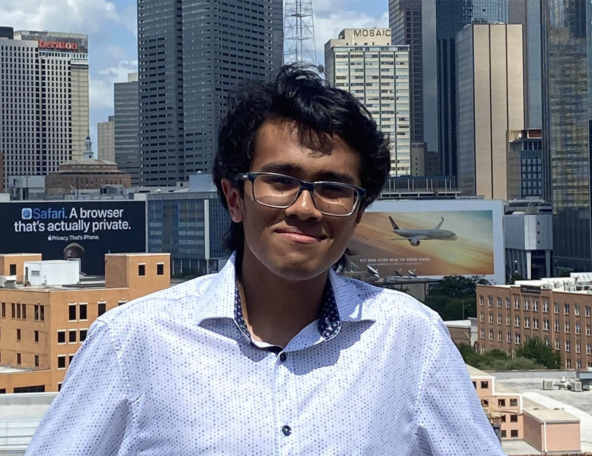 A young man with glasses smiles in front of a city skyline, showcasing tall buildings and a large advertisement featuring an airplane.