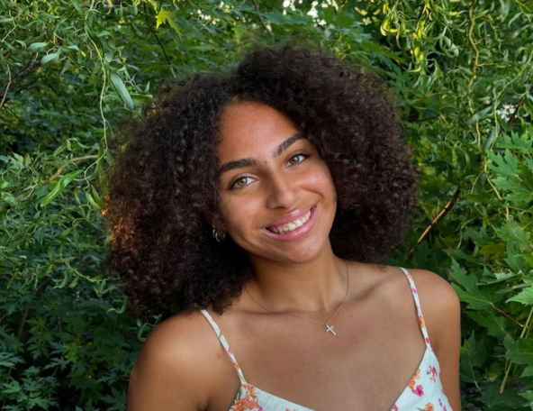 A young woman with curly hair smiles warmly, wearing a floral dress against a backdrop of lush green foliage.