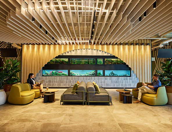 A modern lounge area features two people seated in stylish chairs, an aquarium backdrop, leafy plants, and warm wooden accents.