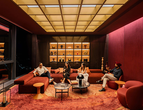 A stylish lounge area features four people seated on a red sectional, surrounded by decorative shelves and warm ambient lighting.